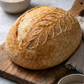 Loaf of bread on a wooden cutting board with bowls of flour and a wooden spoon in the background.