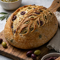 Loaf of bread with olive pieces on a wooden board, accompanied by bowls of olive oil and olive spread.